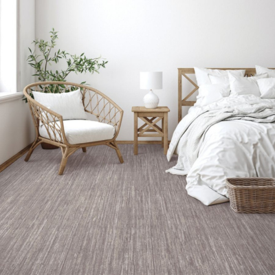 A bright and airy bedroom featuring a light brown and gray striped carpet, a wooden bed with white bedding, a rustic bedside table, a wicker chair, and a leafy green plant by the window.